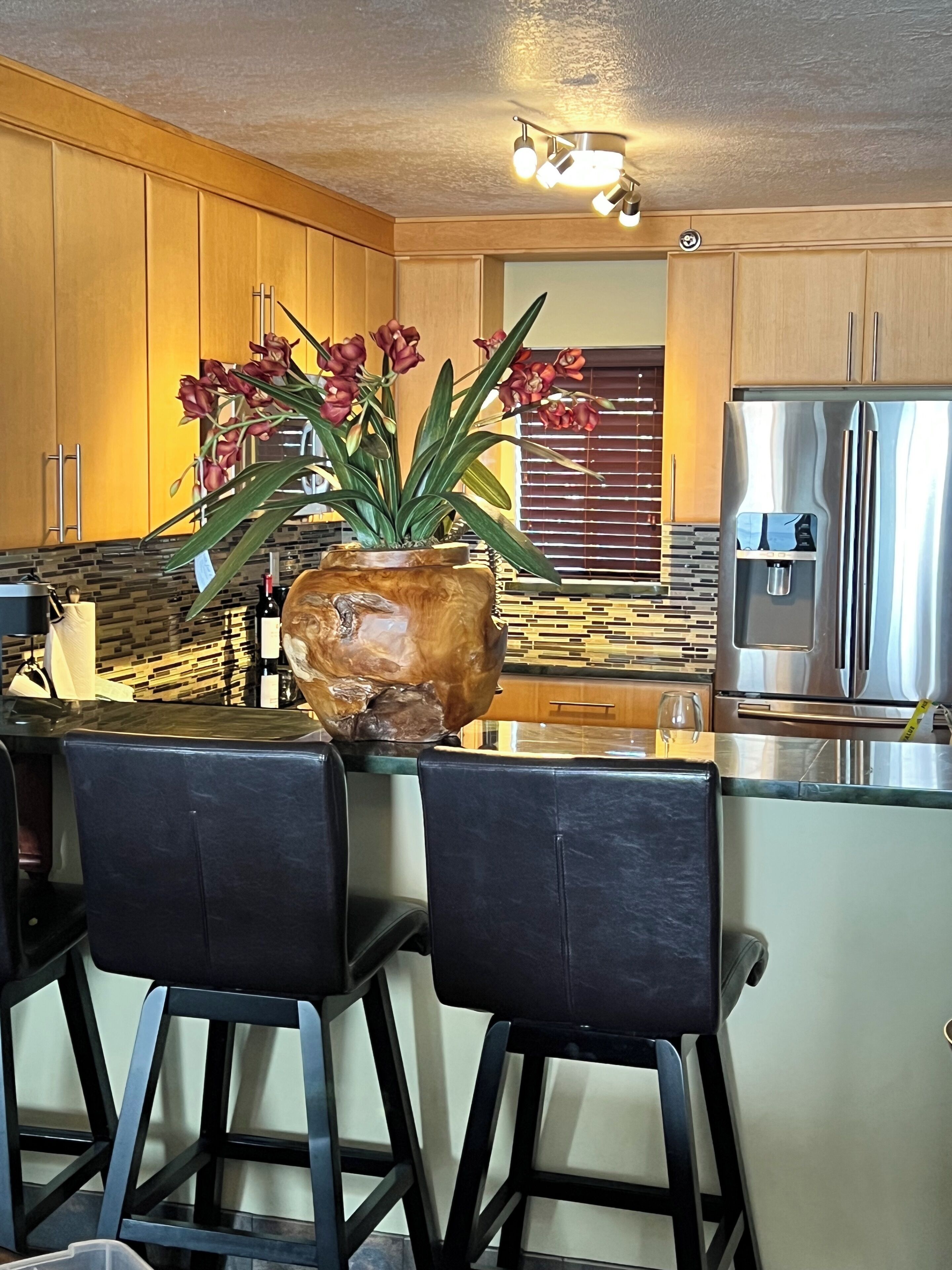 A modern kitchen with wooden cabinets, a stainless steel refrigerator, and a bar area with two black barstools.