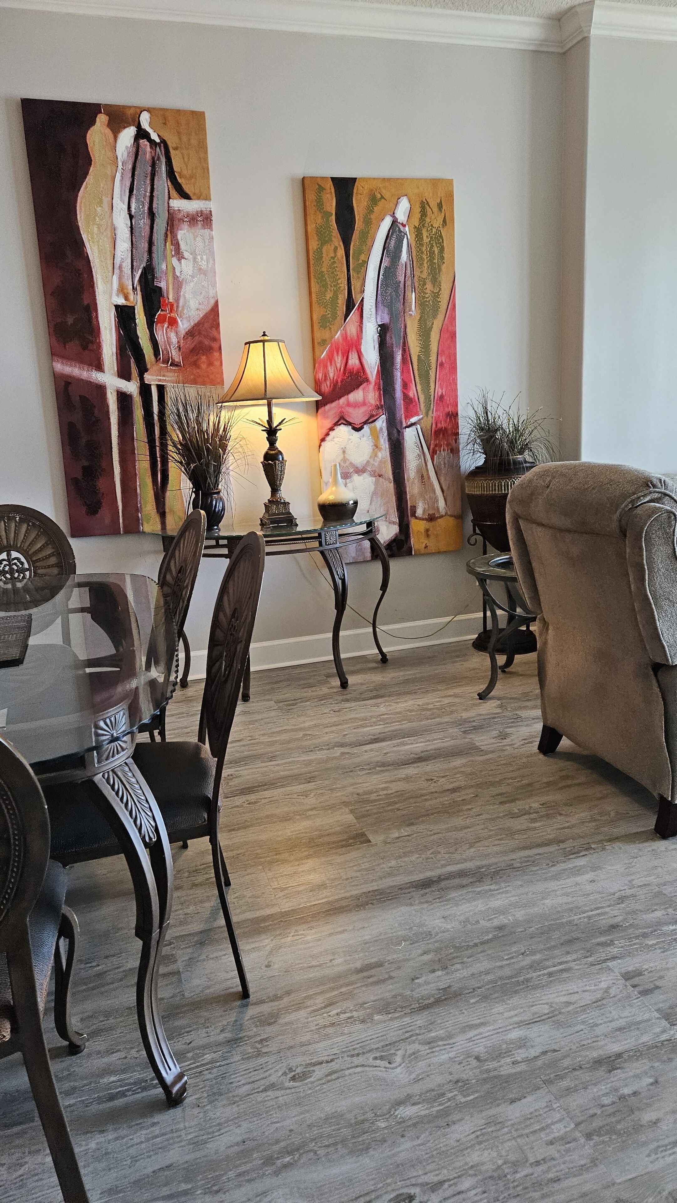 En matplats med ett glasbord, mörka trästolar och en lampa. På väggen hänger två stora tavlor och på golvet står en krukväxt.