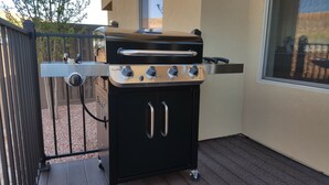 Una parrilla Char-Broil negra con cuatro quemadores y una repisa lateral, ubicada sobre una terraza de madera con barandilla metálica y una ventana al fondo.