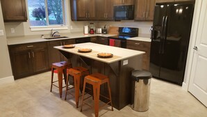 Una cocina con alacenas de madera oscura, un refrigerador negro, un microondas y un lavaplatos. Hay tres taburetes de madera y un bote de basura.