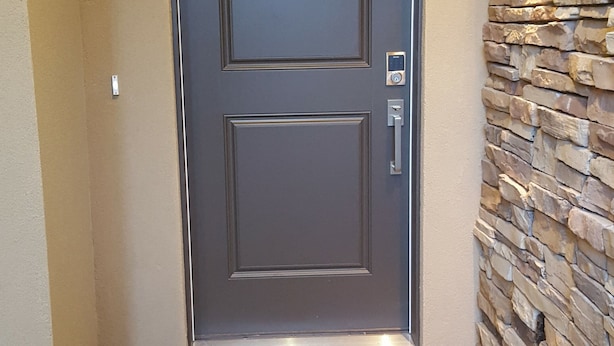 Una puerta oscura con cerradura electrónica y un pequeño panel rectangular, felpudo y pared de piedra con una lámpara.