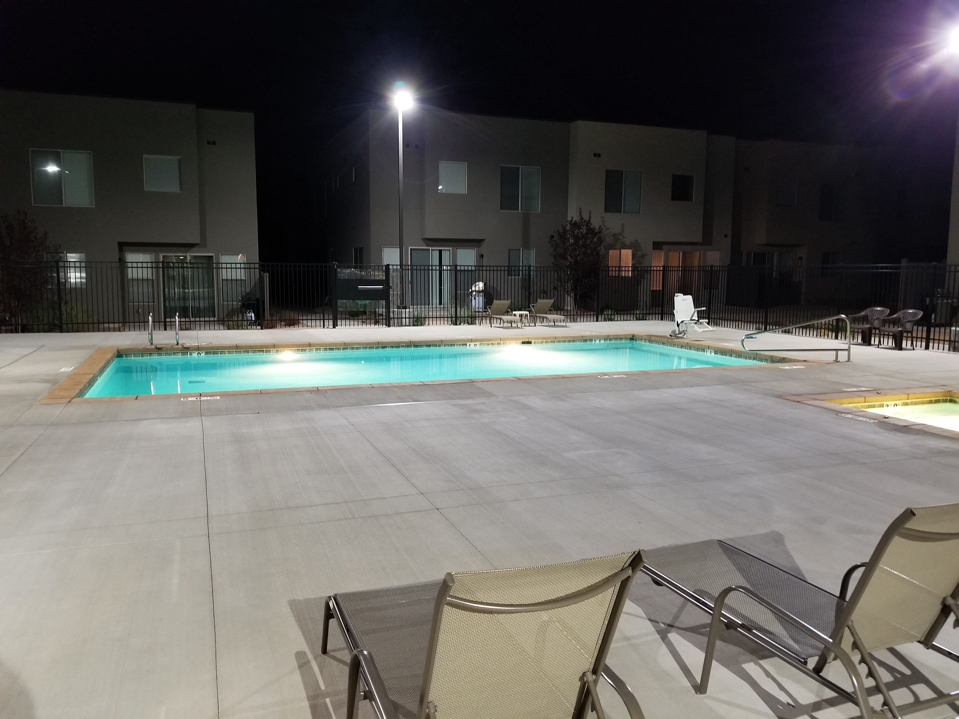 A swimming pool with lounge chairs and a fence, surrounded by buildings at night.