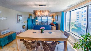 A modern dining area with a wooden table, blue chairs, and a view of the city through large windows. - Surf & Racquet Club A124-Condo (Fernandina Beach)