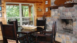 L’intérieur d’un chalet en bois, avec une cheminée en pierre, une table à manger entourée de chaises et une fenêtre donnant sur la verdure.