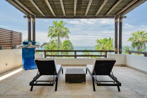Un espace extérieur aménagé, abrité et doté de deux fauteuils confortables et d’un élément décoratif bleu, offrant une vue sur une plage bordée de palmiers et sur l’océan.