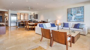A modern living room with a white sofa, wooden chairs, a coffee table, and a dining area with a kitchen in the background. - Beach Front Condo w/ Balcony! Amazing Views, White Sand & Pools! (Punta Mita)