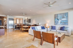 A modern living room with a white sofa, wooden chairs, a coffee table, and a dining area with a kitchen in the background. - Beach Front Condo w/ Balcony! Amazing Views, White Sand & Pools! (Punta Mita)