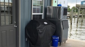 A covered grill and refrigerator on a wooden deck, with a blue bucket nearby.