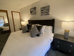 A neatly made bed with white linens and several pillows, a bedside table with a lamp, and two framed pictures on the wall.