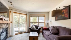 A living room with a fireplace, a sofa, a coffee table, and a large window with blinds. - Ski In Ski Out Great Location Hot Tub Super Clean Fully Stocked Great Reviews (Whistler)