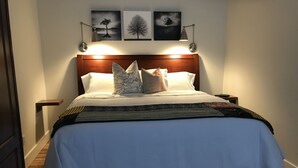 Un lit bien fait, doté d’une tête de lit en bois, flanqué de tables de chevet avec des lampes, et trois œuvres d’art encadrées au-dessus de la tête de lit.