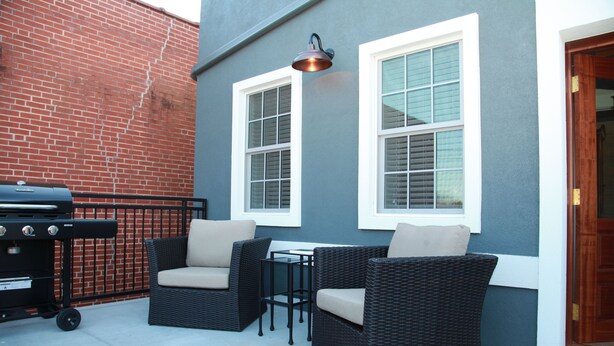 Une terrasse extérieure avec des meubles en osier, un barbecue et un mur percé de deux fenêtres.