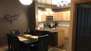 A modern kitchen with wooden cabinets, a microwave, and a stove. There is a dining area with a black table and chairs, and a small island with a sink.
