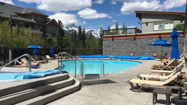 A pool area with lounge chairs and umbrellas, surrounded by buildings and trees, with a mountain in the background.