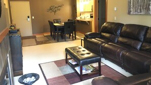A living room with a leather sofa, a coffee table, a dining table with chairs, a kitchen area, and a door leading to another room.