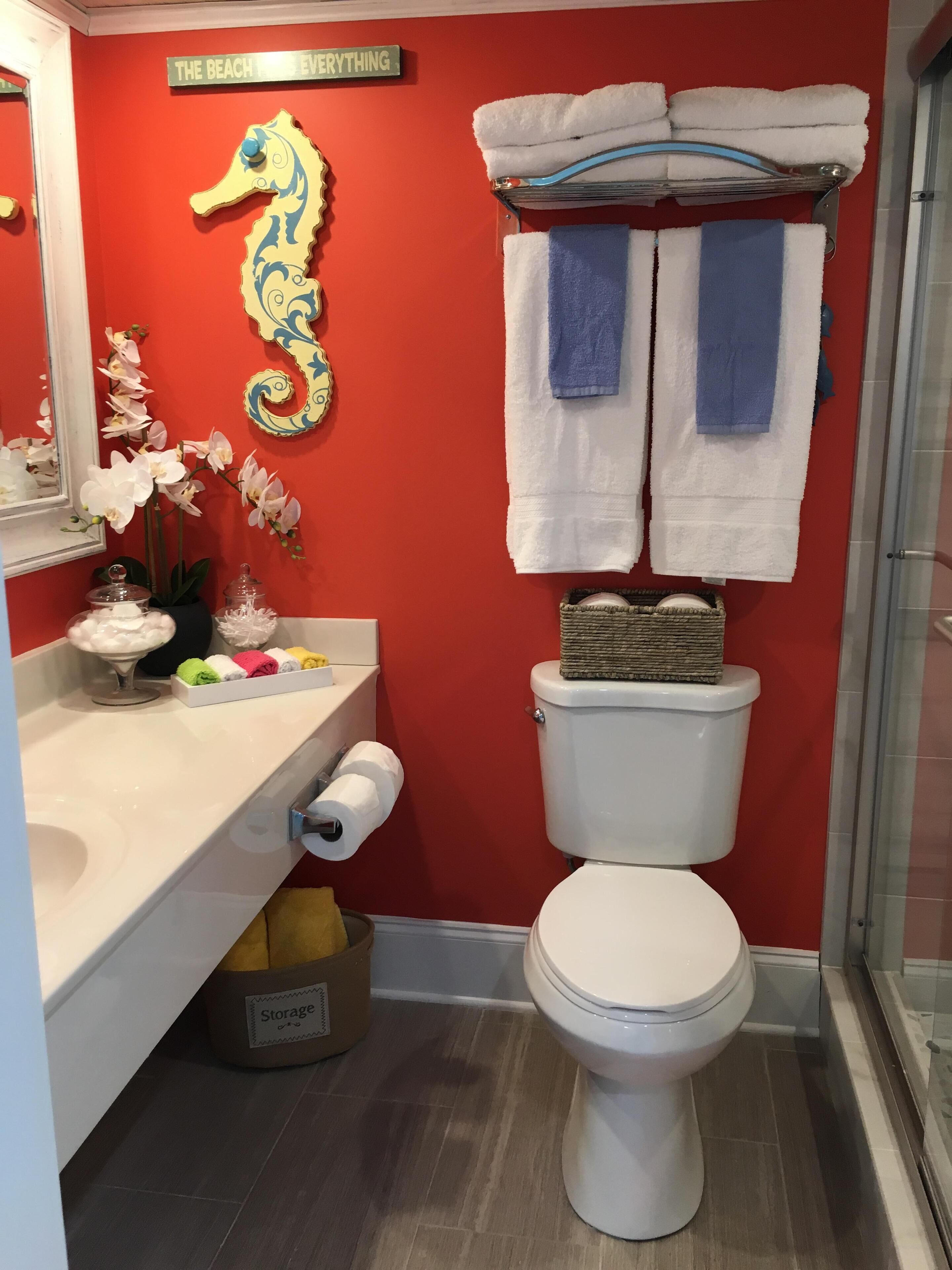 Een badkamer met een rode wand, een decoratie van een zeepaard, een handdoekhouder met handdoeken, een toilet, een wastafel met zeepdispenser en een opbergbak.