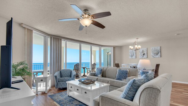 A spacious living room with a ceiling fan, a large window with a view of the ocean, a white coffee table, and a sofa with blue pillows. - Long Beach 2-Bedroom Condo with Two Seasonal Beach Chairs with Umbrella (Panama City Beach)