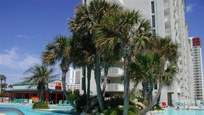 A swimming pool with palm trees and lounge chairs, surrounded by a high-rise building. - Long Beach 2-Bedroom Condo with Two Seasonal Beach Chairs with Umbrella (Panama City Beach)