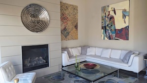 A modern living room with a fireplace, a white sofa, a glass coffee table with decorative items, and three artworks on the walls. - Palm Springs - Central & Modern! (Palm Springs)
