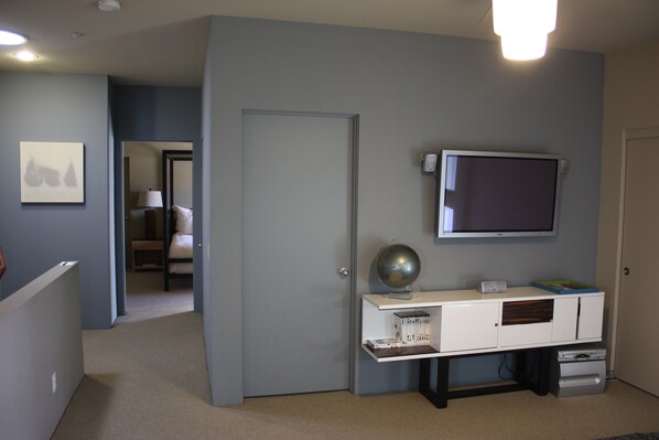 A room with a flat-screen TV mounted on the wall, a white console table with a globe and books, and a door leading to another room. - Palm Springs - Central & Modern! (Palm Springs)