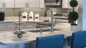Une cuisine moderne avec des meubles blancs, un évier en acier inoxydable, un four à micro-ondes et une cuisinière. On y trouve deux tabourets de bar bleus et une petite plante en pot.