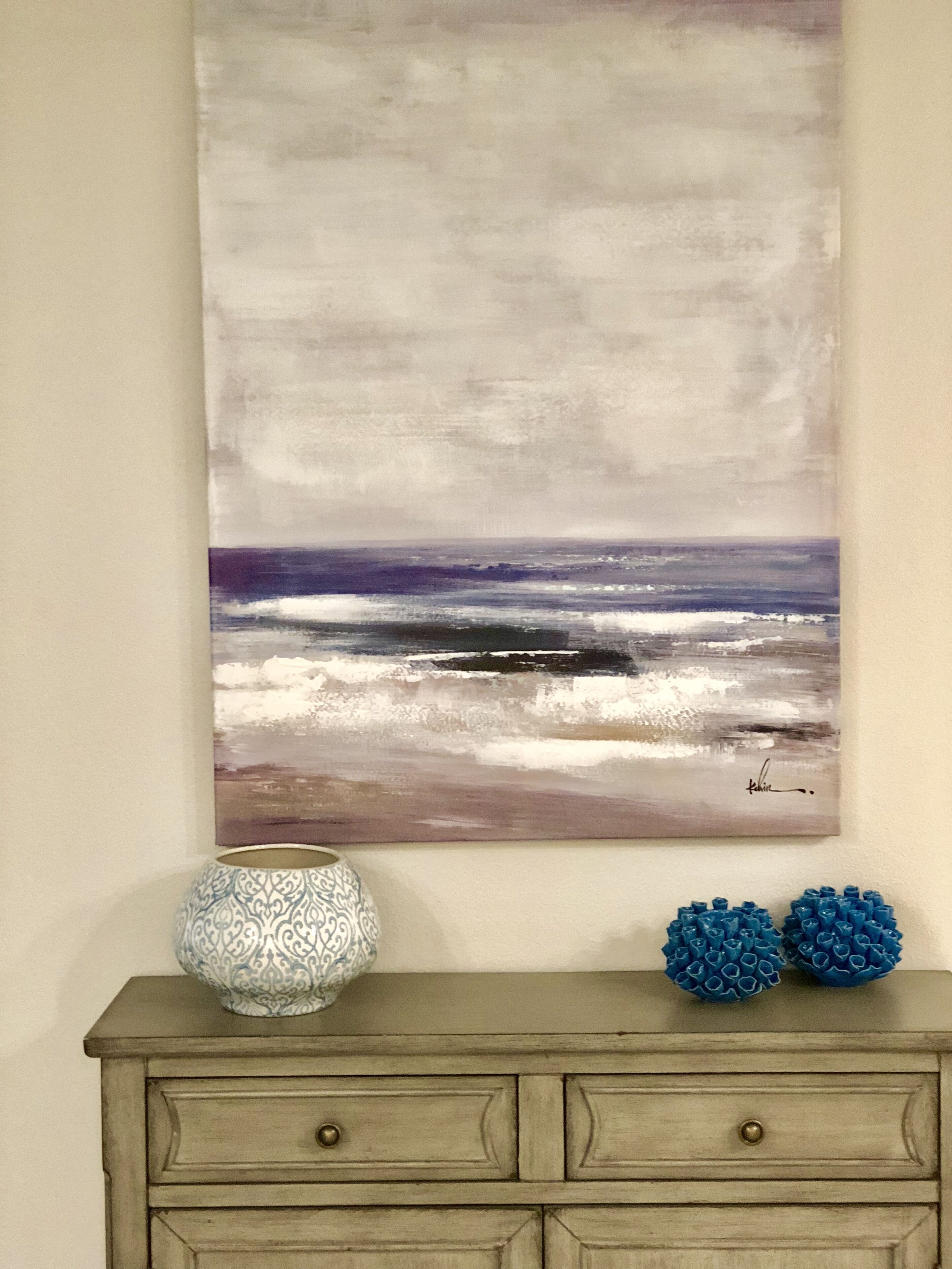 A wall-mounted abstract painting of a beach scene, a patterned vase, and decorative blue spheres on a wooden dresser.