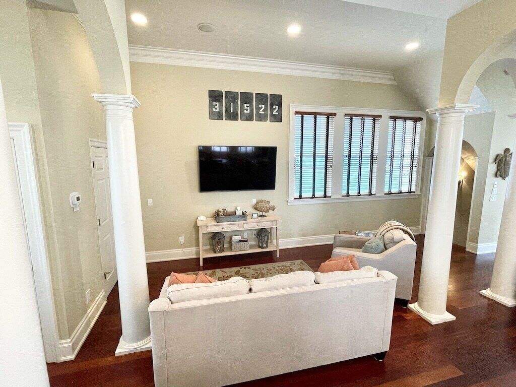Uma sala de estar com um sofá branco, uma TV de tela plana na parede e uma janela com persianas.