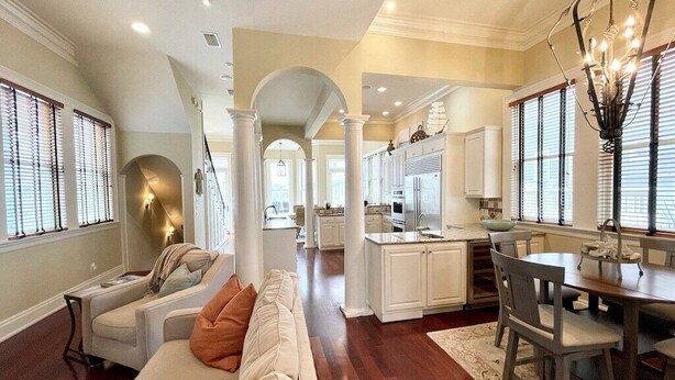 Uma sala de estar ampla com um sofá grande, um espaço de refeições com mesa e cadeiras, e uma cozinha com armários brancos e um lustre.