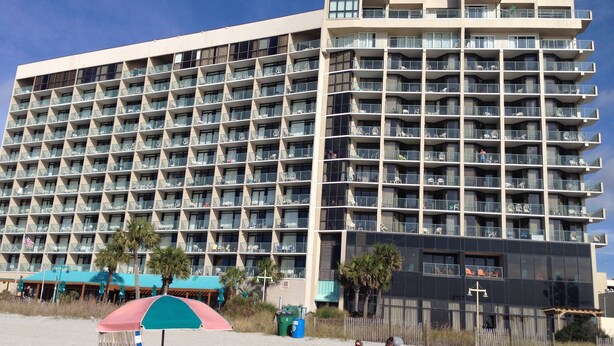 A large hotel with multiple balconies and a beach area with umbrellas and lounge chairs.