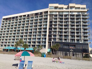 A large hotel with multiple balconies and a beach area with umbrellas and lounge chairs.