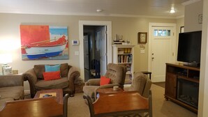 Ein Wohnzimmer mit einer Couch, Sesseln, einem Couchtisch, einem Bücherregal, einem Fernseher und einem Bild eines Bootes.