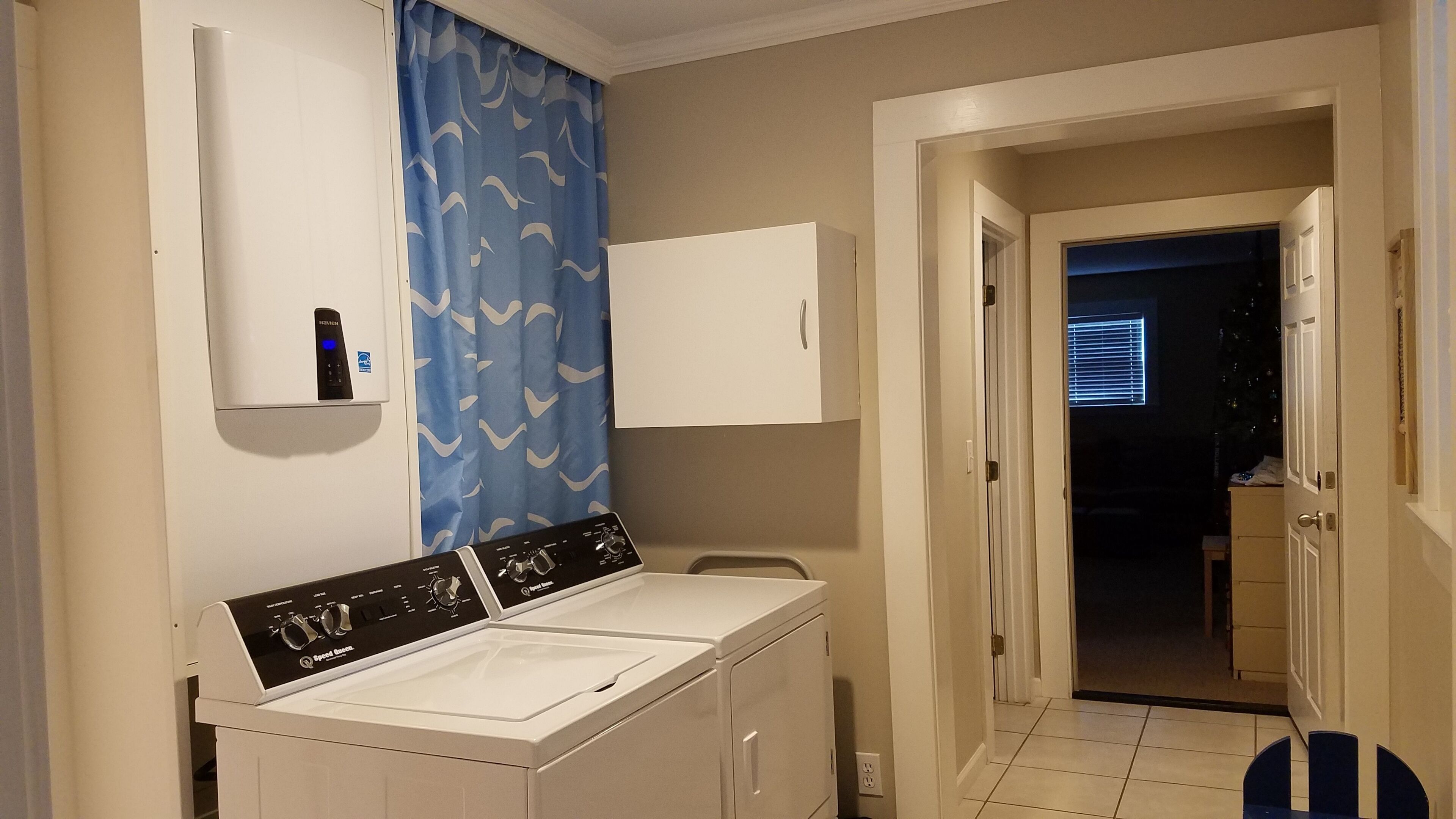 Un espace buanderie comprenant un lave-linge et un sèche-linge à chargement frontal, un meuble de rangement au-dessus du sèche-linge et un rideau bleu orné d’un motif ondulé.