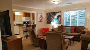 Ein Wohnzimmer mit einer Couch, Sesseln, einem Couchtisch und einem Fernseher. Es gibt eine Küchenzeile mit einer Mikrowelle und einem Spülbecken.