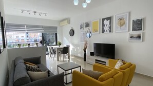 Ein modernes Wohnzimmer mit einer Couch, gelben Sessel, einem Fernseher und einer Essecke mit Tisch und Stühlen.