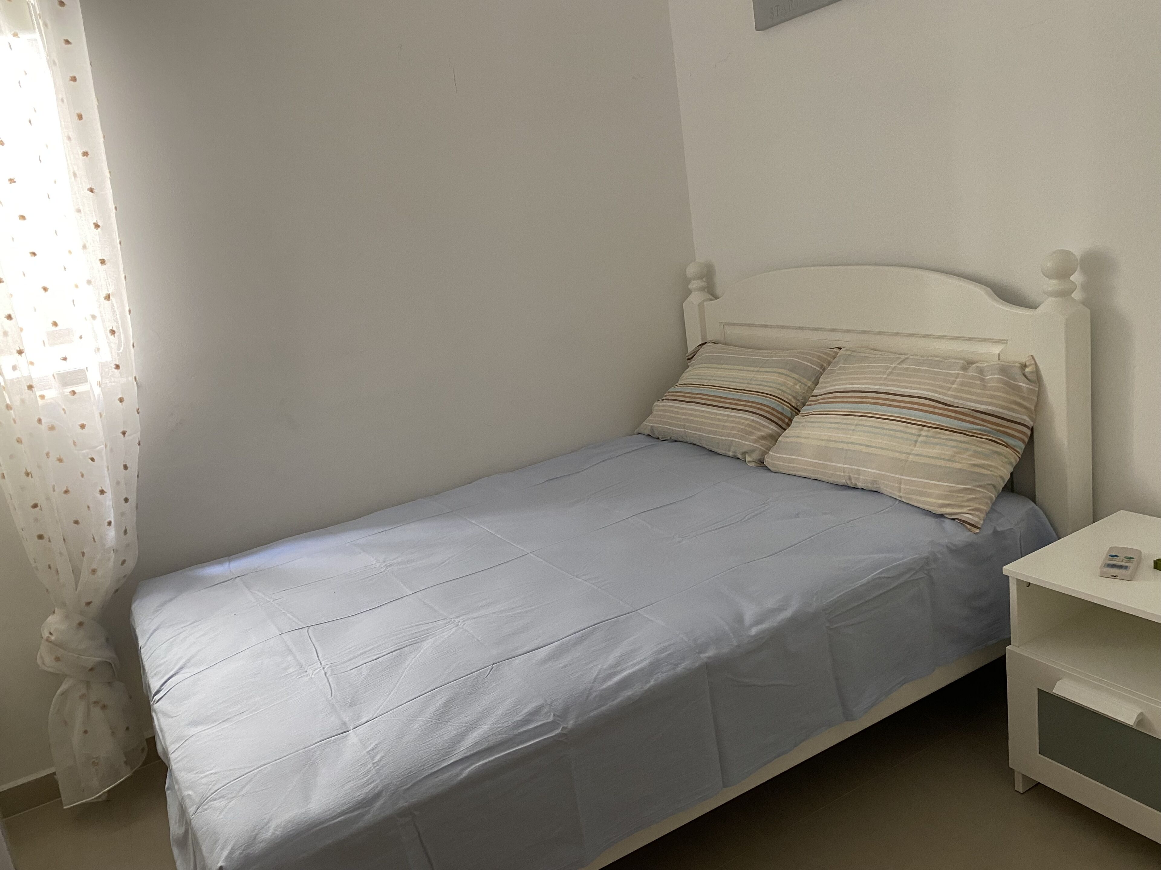 A small, neatly made bed with a light blue bedspread and two striped pillows, a white nightstand with a small lamp, and a sheer curtain with a floral pattern.
