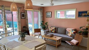 A cozy living room with a sofa, a dining table, and chairs. There are plants, a coffee table, and a window with curtains.