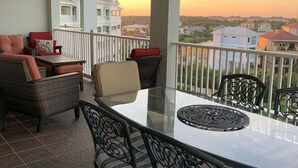 Un balcón con una mesa y sillas de vidrio, vistas al vecindario y un atardecer de fondo.