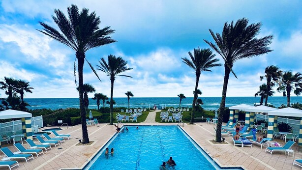Una piscina con sillas de descanso y palmeras, con vista al océano de fondo.