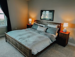A bedroom with a wooden bed, two bedside tables with lamps, a window with curtains, and a framed picture on the wall.