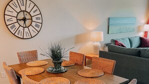 A dining area with a wooden table, wicker chairs, a wall clock, and a blue painting on the wall.