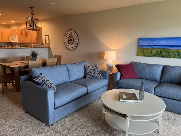A living room with two sofas, a coffee table, a dining area with a table and chairs, a kitchen with wooden cabinets, a clock, and a painting on the wall.
