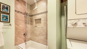 A bathroom with a glass-enclosed shower, a bathtub, and a washing machine. - Lovely Two Bedroom that Feels Like Home (Waikoloa)