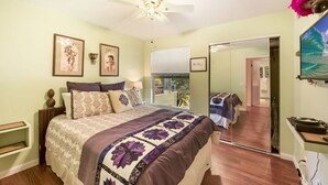 A bedroom with a bed, a ceiling fan, a window, a mirror, and a door leading to another room. - Lovely Two Bedroom that Feels Like Home (Waikoloa)