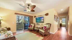 A spacious living room with a ceiling fan, a large window, and a view of the outdoors. - Lovely Two Bedroom that Feels Like Home (Waikoloa)