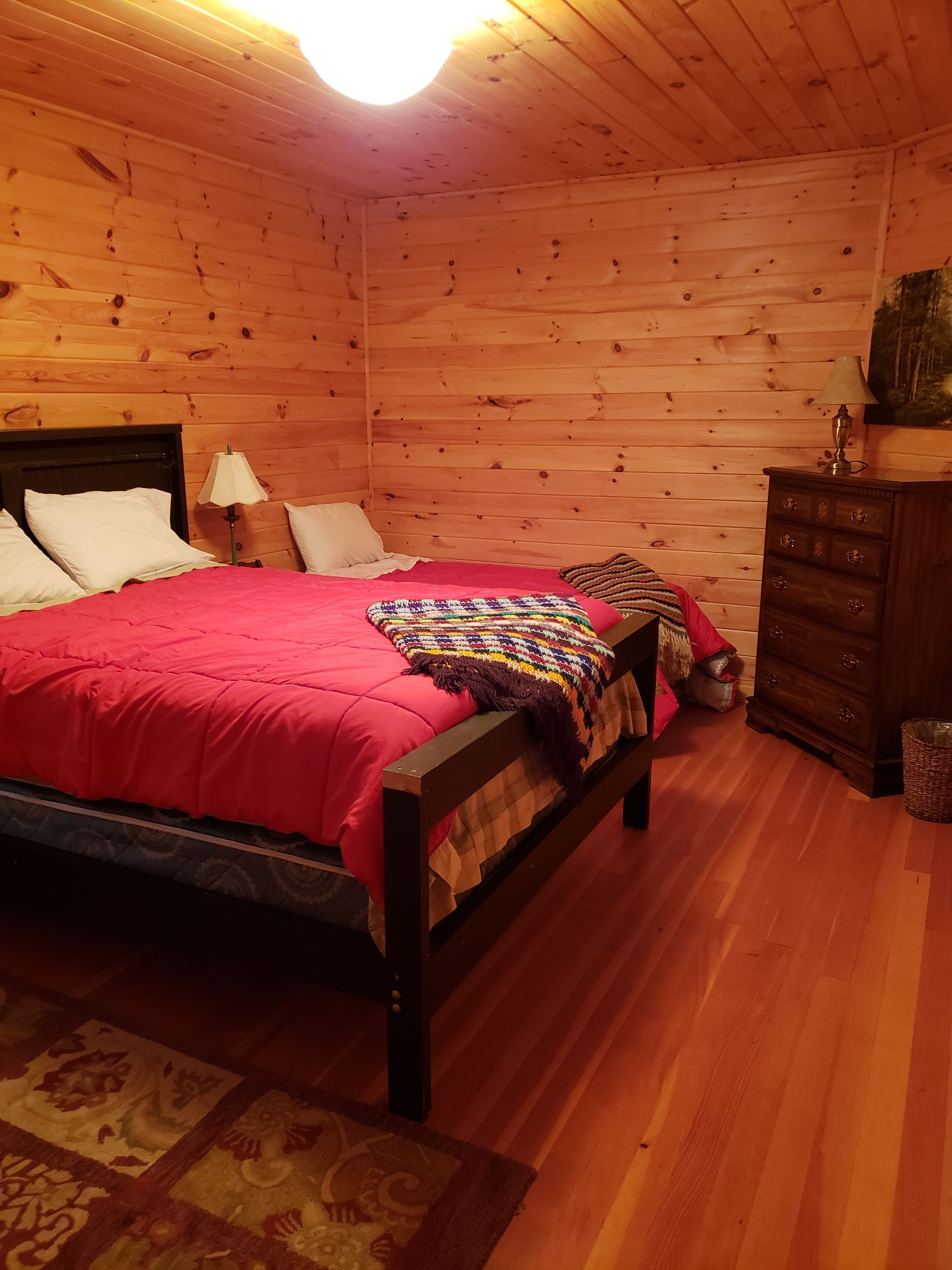 Een slaapkamer met houten lambrisering, een bed, kussens, een lamp, een dressoir en een vloerkleed.