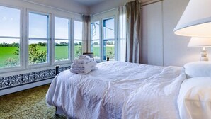 Ein Hotelzimmer mit einem großen Bett, einer Kandelaber, einer Lampe und Blick auf ein grünes Feld durch große Fenster.
