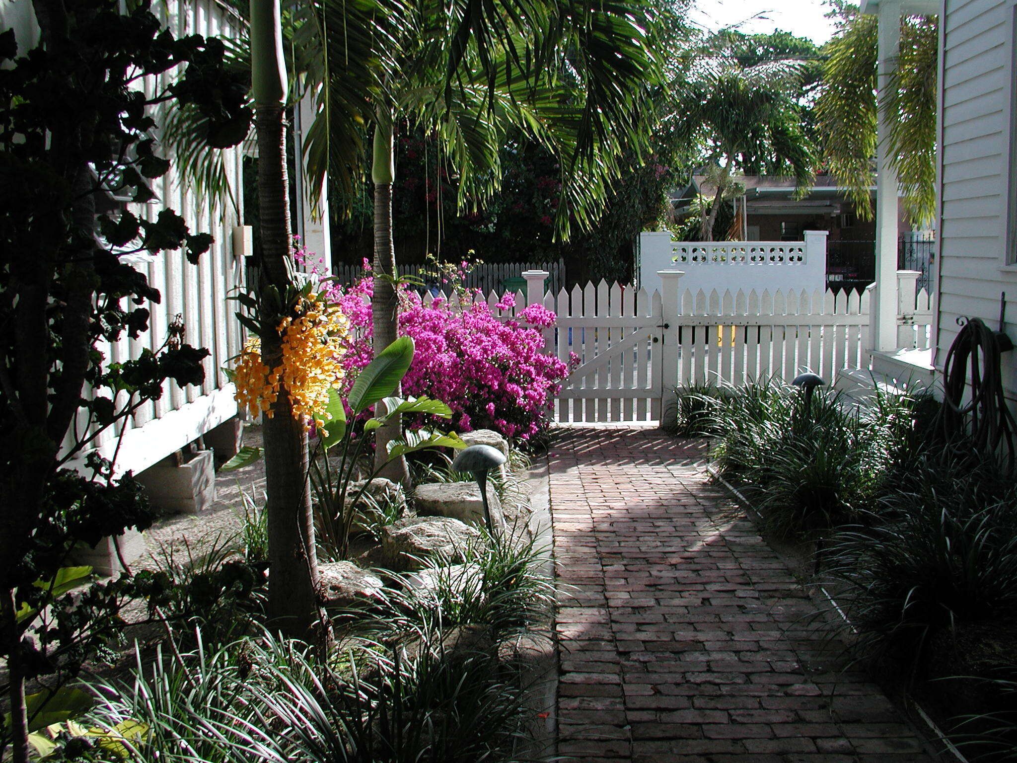 Un sendero en un jardín conduce a una cerca blanca con una puerta, rodeado de exuberante vegetación tropical y flores.