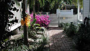 Ein Gartenweg führt zu einem weißen Zaun mit einem Tor, umgeben von üppigen tropischen Pflanzen und Blumen.