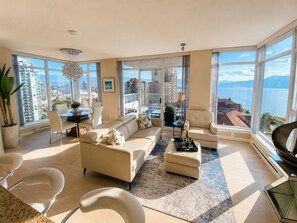 A spacious living room with a large sofa, a dining area, and a view of the city and sea. - The Skye at Waterscapes Kelowna Luxurious Lake View Condo (Kelowna)