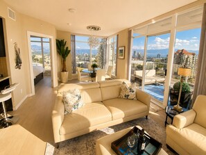 A spacious living room with a large sofa, a dining area, and a view of the cityscape through large windows. - The Skye at Waterscapes Kelowna Luxurious Lake View Condo (Kelowna)
